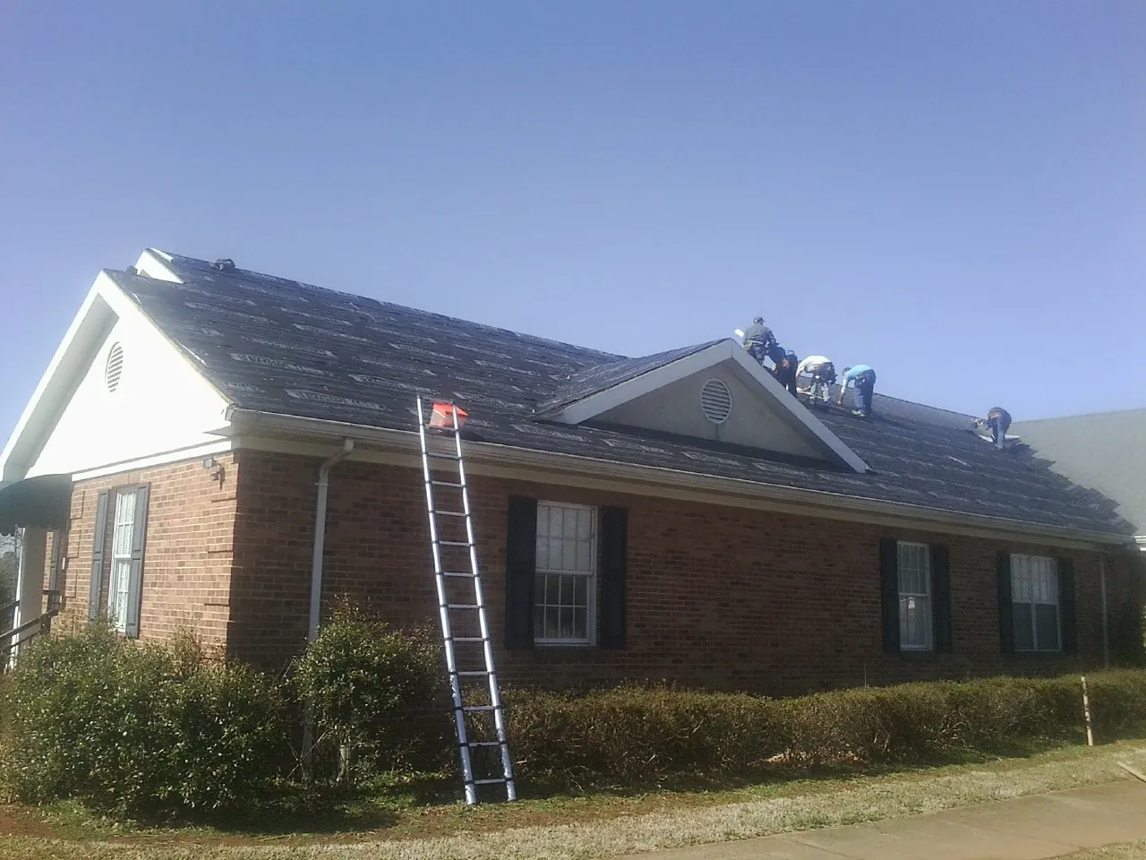 Crew tearing shingles off a brick ranch roof with a ladder set — Emergency Roof Repair work by our crew in Centereach