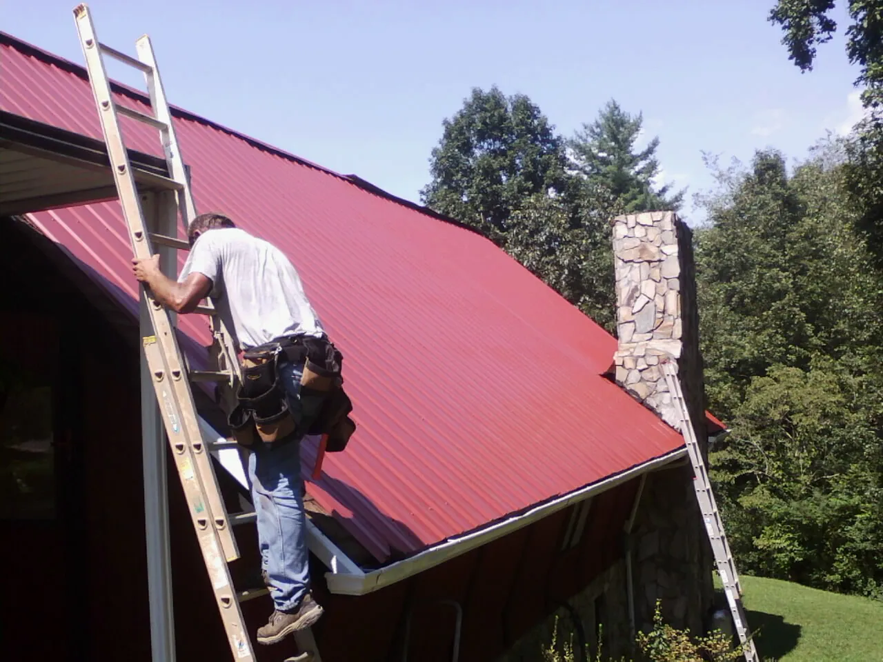 Roofer on a ladder beside a red metal roof and stone chimney — Soffit & Fascia Repair by Roofing Brothers in Centereach
